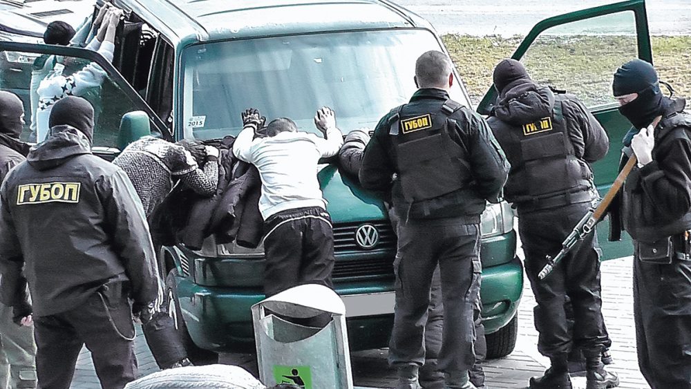 Силовики в форме ГУБОПиК задерживают людей у микроавтобуса, задержанные стоят с поднятыми руками, прижатыми к машине.